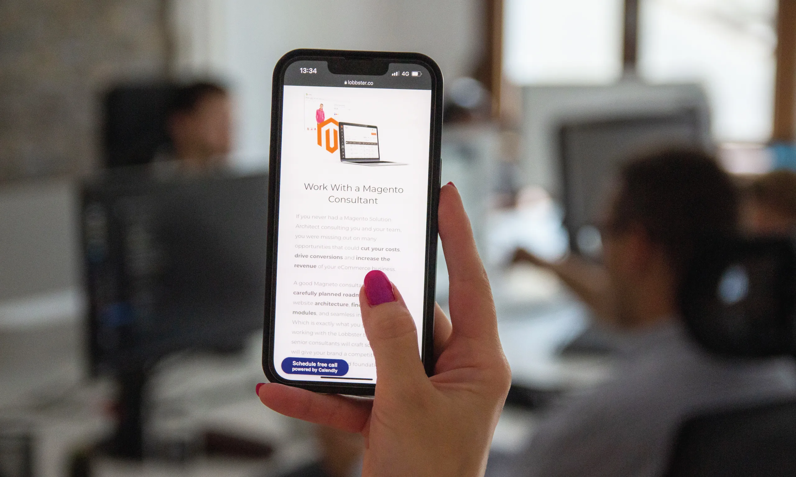 A close up of a mobile phone with the text about B2B Magento consulting services. Some bleary people working in the background.